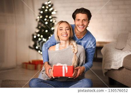 Lovely couple with gift in front of Christmas tree smiling to camera, sitting on floor 130576052