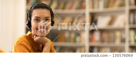 E-teaching concept. Portrait of happy woman in headset sitting in library and smiling at camera, copy space. Online teaching, modern distance education and remote e-learning, copy space 130576069