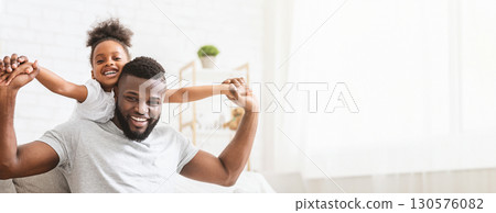 A delighted black father shares a playful moment with his young daughter in a well-lit living room. The little girl sits on her father shoulders, copy space A delighted black father shares a playful moment with his young daughter in a well-lit living room. The little girl sits on her father shoulders, copy space 130576082