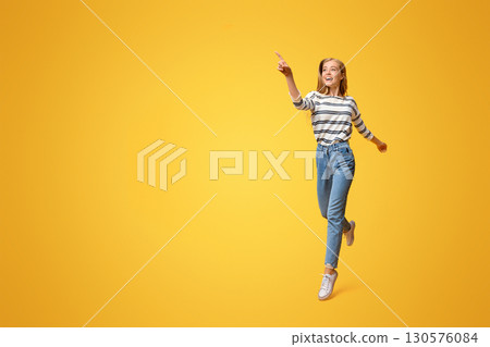 Look there. Joyful girl jumping and pointing on copy space, showing something on yellow background Look there. Joyful girl jumping and pointing on copy space, showing something on yellow background 130576084