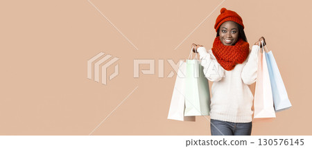 A joyful young African American woman stands smiling while holding several colorful shopping bags. She is dressed warmly in a cozy sweater and a bright red winter hat, copy space 130576145