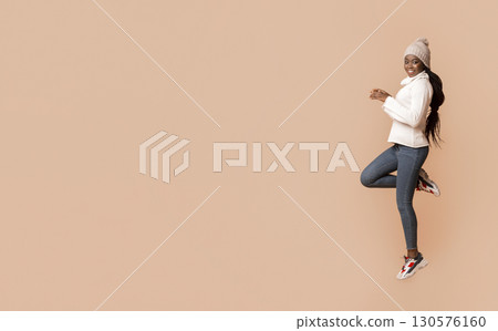Sporty winter african american woman jumping on one leg over beige background, winter activity concept Sporty winter african american woman jumping on one leg over beige background, winter activity concept 130576160