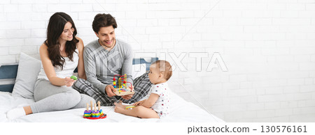 A young couple enjoys quality time together in a bright room while engaging their baby with colorful toys on a comfortable bed. The atmosphere is filled with warmth and joy, copy space A young couple enjoys quality time together in a bright room while engaging their baby with colorful toys on a comfortable bed. The atmosphere is filled with warmth and joy, copy space 130576161