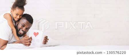 A joyful black father and daughter share a warm moment together while displaying a heart drawing created by the girl. They sit on a bed, with smiles lighting up their faces, copy space A joyful black father and daughter share a warm moment together while displaying a heart drawing created by the girl. They sit on a bed, with smiles lighting up their faces, copy space 130576220