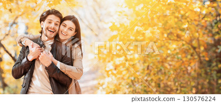 Positive woman hugging her happy man from behind, posing at autumn park, copy space Positive woman hugging her happy man from behind, posing at autumn park, copy space 130576224