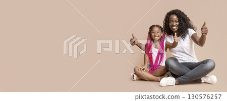Family approval. Black Mother and Daughter Gesturing Thumbs Up Sitting On Floor Over Yellow Studio Background With Copy Space 130576257