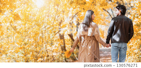 Rear view of young man and woman lovers holding hands and walking by golden forest, sun flare, copy space 130576271