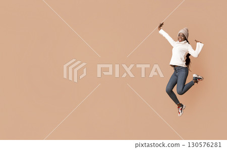 Lifeful black woman in warm hat enjoying winter over beige studio background, jumping up, copy space Lifeful black woman in warm hat enjoying winter over beige studio background, jumping up, copy space 130576281