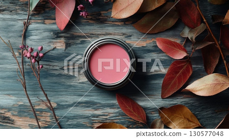 A blush compact sits centered on a weathered wooden surface, framed by vibrant autumn leaves in shades of red and orange. Small pink flowers complement the earthy tones, creating a serene setting. A blush compact sits centered on a weathered wooden surface, framed by vibrant autumn leaves in shades of red and orange. Small pink flowers complement the earthy tones, creating a serene setting. 130577048