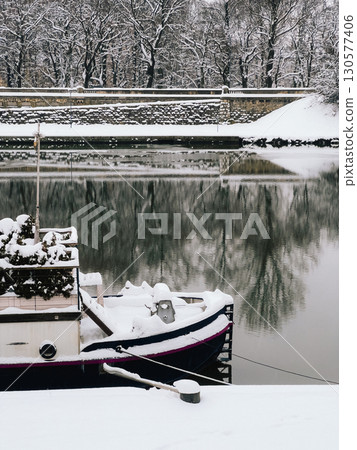Tranquil winter scene features houseboat covered in snow, moored on calm river reflecting snowy trees and stone embankment, creating serene and picturesque winter landscape. Snowcovered houseboat 130577406