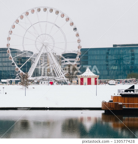 Ferris wheel and carousel standing still in snow covered park during winter next to modern glass buildings with river in foreground, in Krakow, Poland. Landscape reveal snow blanketed Krakow park 130577428