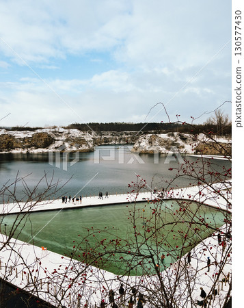 Tourists are walking near an artificial body of water created in former quarry, during winter, with snow covering ground and red berries on branches. Tourists walking near flooded limestone quarry Tourists are walking near an artificial body of water created in former quarry, during winter, with snow covering ground and red berries on branches. Tourists walking near flooded limestone quarry 130577430
