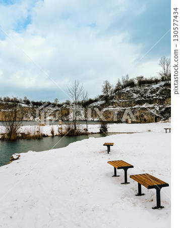 Wooden benches standing on snowy shore overlooking turquoise waters, surrounded by cliffs and vegetation, during cold winter day. Wooden benches resting on snow covered shore, rocky cliffs, winter Wooden benches standing on snowy shore overlooking turquoise waters, surrounded by cliffs and vegetation, during cold winter day. Wooden benches resting on snow covered shore, rocky cliffs, winter 130577434