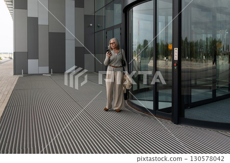 Older woman using smartphone while waiting outside modern building in urban area 130578042
