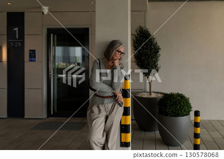 Senior woman leaning against a post while waiting outside a modern building during daylight hours 130578068