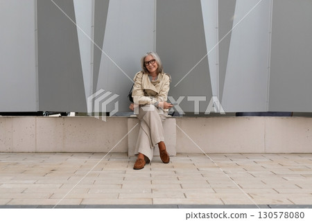 Senior woman sits casually against modern building backdrop in an urban setting during daytime 130578080