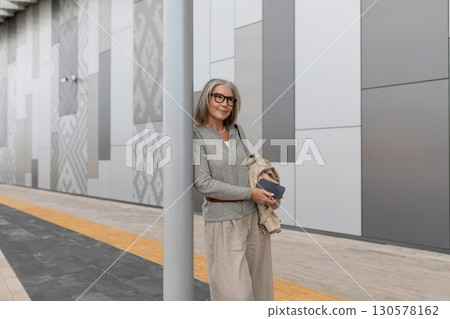 Confident woman stands by modern urban wall while holding phone and jacket in late afternoon 130578162