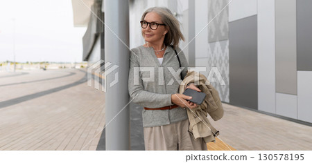 Mature woman enjoying a stroll by modern architecture on a cloudy day while holding a phone and coat 130578195