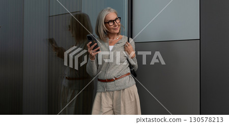 Woman with glasses checks her phone while standing against a modern building wall in an urban 130578213