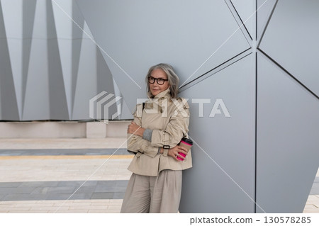 Stylish older woman leans against modern architecture while enjoying a coffee in an urban setting Stylish older woman leans against modern architecture while enjoying a coffee in an urban setting 130578285