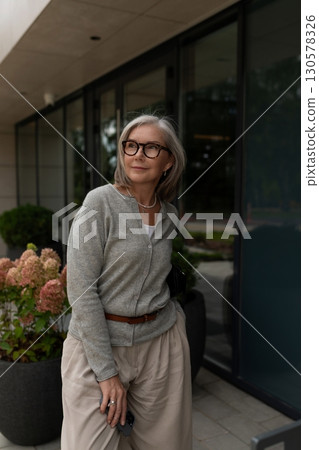 Elderly woman in stylish outfit enjoying a sunny day outside modern building with plants Elderly woman in stylish outfit enjoying a sunny day outside modern building with plants 130578326