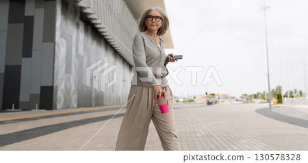Woman walking confidently outside a modern building while holding a pink reusable cup and listening 130578328