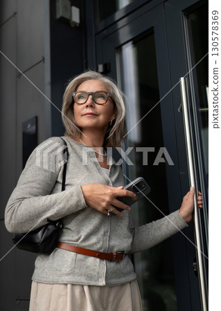 Senior woman entering a modern building while holding a smartphone in an urban setting Senior woman entering a modern building while holding a smartphone in an urban setting 130578369