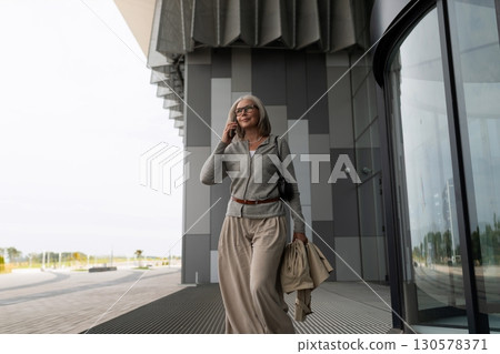 Senior woman leaving modern building while talking on smartphone in urban setting during daylight Senior woman leaving modern building while talking on smartphone in urban setting during daylight 130578371