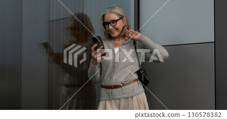 Woman in cozy sweater smiles while using smartphone near modern building on sunny day Woman in cozy sweater smiles while using smartphone near modern building on sunny day 130578382