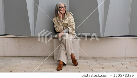Stylish older woman sits thoughtfully against modern architecture in urban setting during daylight Stylish older woman sits thoughtfully against modern architecture in urban setting during daylight 130578389