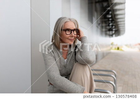 Woman in gray sweater relaxes outdoors while thinking softly next to modern architecture during 130578408
