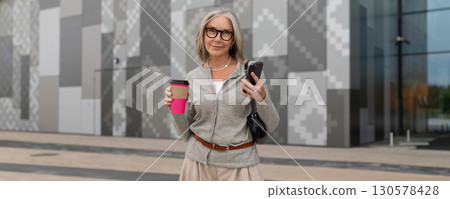 Woman holding coffee and smartphone while standing in modern urban setting during daytime 130578428