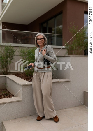 Middle-aged woman posing outdoors in fashionable attire near modern architecture on a sunny day 130578444
