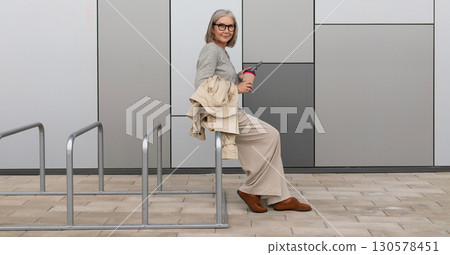 Older woman sitting casually on bike rack while holding a beverage near modern building during 130578451