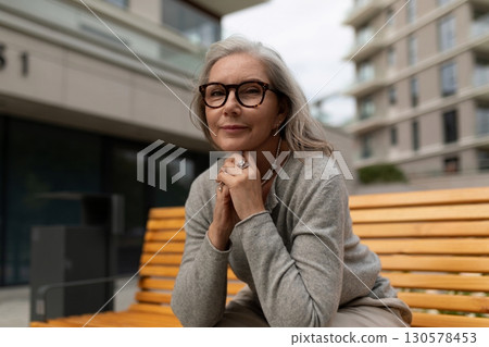 Senior woman enjoys a moment of reflection on a bench in a modern urban setting during a cloudy 130578453