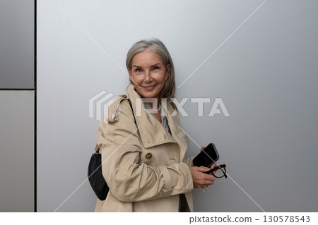 Smiling woman in a beige trench coat holds glasses and smartphone against a modern background in an 130578543