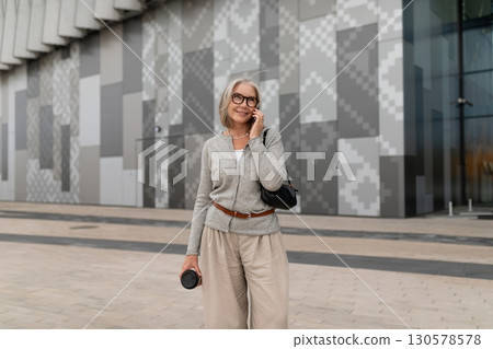 Smiling senior woman enjoying a phone call outside a modern building in the afternoon 130578578