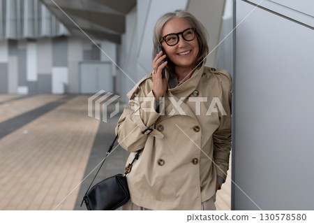 Woman in a beige coat talking on the phone while standing near a modern building in the city during Woman in a beige coat talking on the phone while standing near a modern building in the city during 130578580