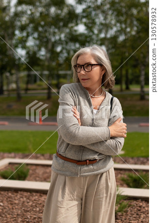 Mature woman enjoys a sunny day while standing outside in a park surrounded by green trees 130578932