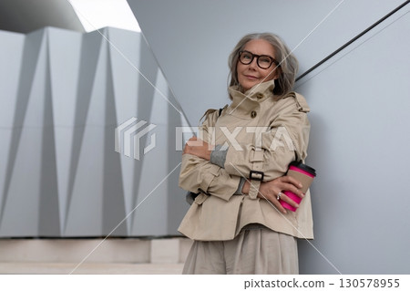 Stylish older woman stands confidently with coffee in modern architectural setting on a bright day Stylish older woman stands confidently with coffee in modern architectural setting on a bright day 130578955