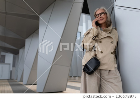 Older woman wearing trench coat talks on phone near modern architecture in urban setting 130578973