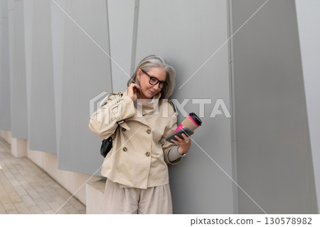 Older woman in beige coat holding coffee cup while checking phone against modern gray wall Older woman in beige coat holding coffee cup while checking phone against modern gray wall 130578982