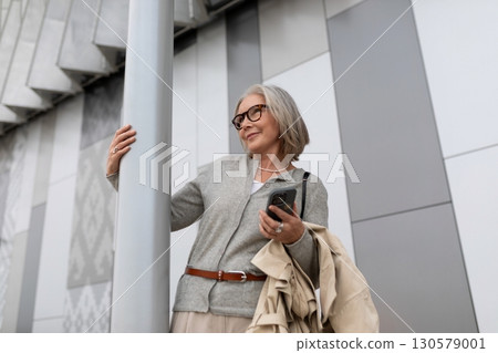 Senior woman with smartphone poses near modern architecture during a sunny afternoon Senior woman with smartphone poses near modern architecture during a sunny afternoon 130579001