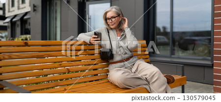 Woman sitting on wooden bench outside modern building while using smartphone during cloudy day 130579045