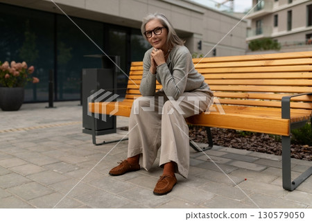 Elderly woman sitting comfortably on a wooden bench in a modern outdoor setting during the day Elderly woman sitting comfortably on a wooden bench in a modern outdoor setting during the day 130579050
