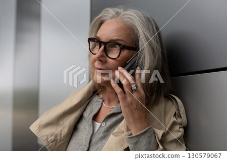 Woman speaking on mobile phone while resting against modern surface in urban setting during daylight Woman speaking on mobile phone while resting against modern surface in urban setting during daylight 130579067