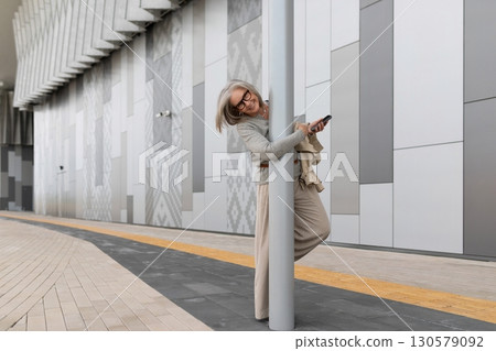 Smiling woman enjoying her smartphone while playfully leaning on a pole in a modern urban setting Smiling woman enjoying her smartphone while playfully leaning on a pole in a modern urban setting 130579092