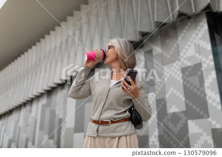 Woman enjoying a coffee while standing outside a modern building showcasing geometric patterns Woman enjoying a coffee while standing outside a modern building showcasing geometric patterns 130579096