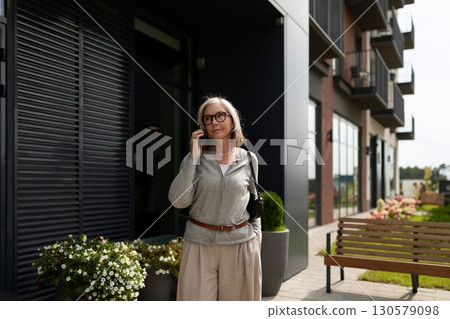 Mature woman checking her phone while standing outside a modern building in a serene urban setting 130579098