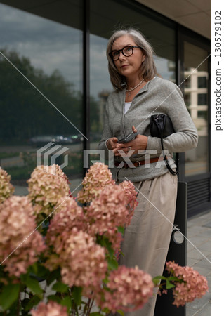 Stylish woman stands near blooming flowers outside a modern building on a cloudy day Stylish woman stands near blooming flowers outside a modern building on a cloudy day 130579102